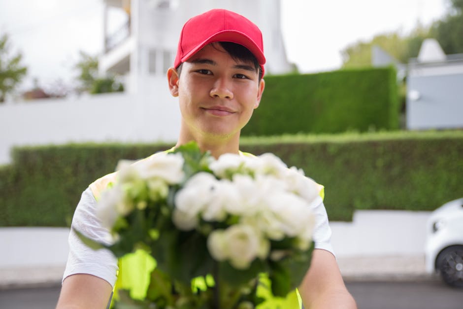 A delivery person wearing a red cap and high-visibility vest is holding a bouquet of fresh white flowers, including roses and possibly hydrangeas, wrapped in green paper. The flowers are vibrant and w