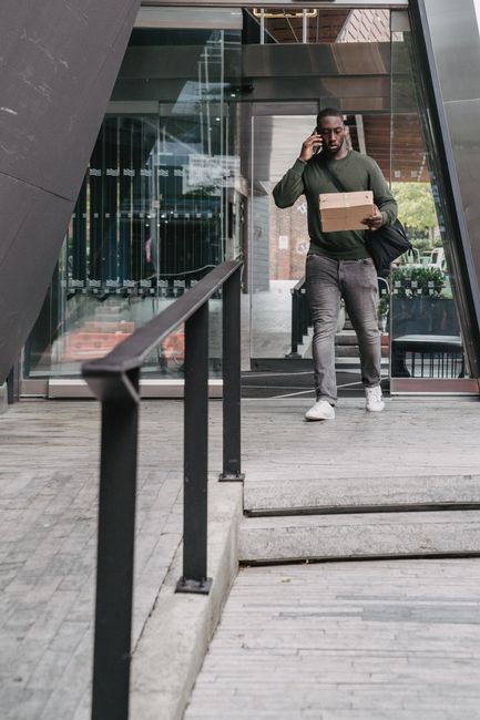A person standing outside a modern glass building, holding a cardboard box in one hand and talking on a mobile phone with the other. The individual is dressed casually in a green sweater, grey jeans,