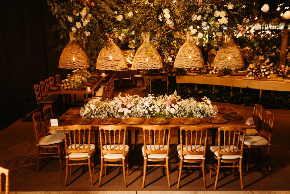 A large, rectangular wooden dining table set for an event, decorated with a lush, elongated floral arrangement consisting of white roses, baby's breath, and soft pink blooms. The table is surrounded b