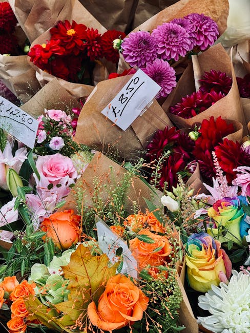 A vibrant assortment of fresh flowers displayed in brown paper wrapping, including bright red dahlias, purple chrysanthemums, pink and peach roses, and multi-colored rainbow roses. The bouquets are ar