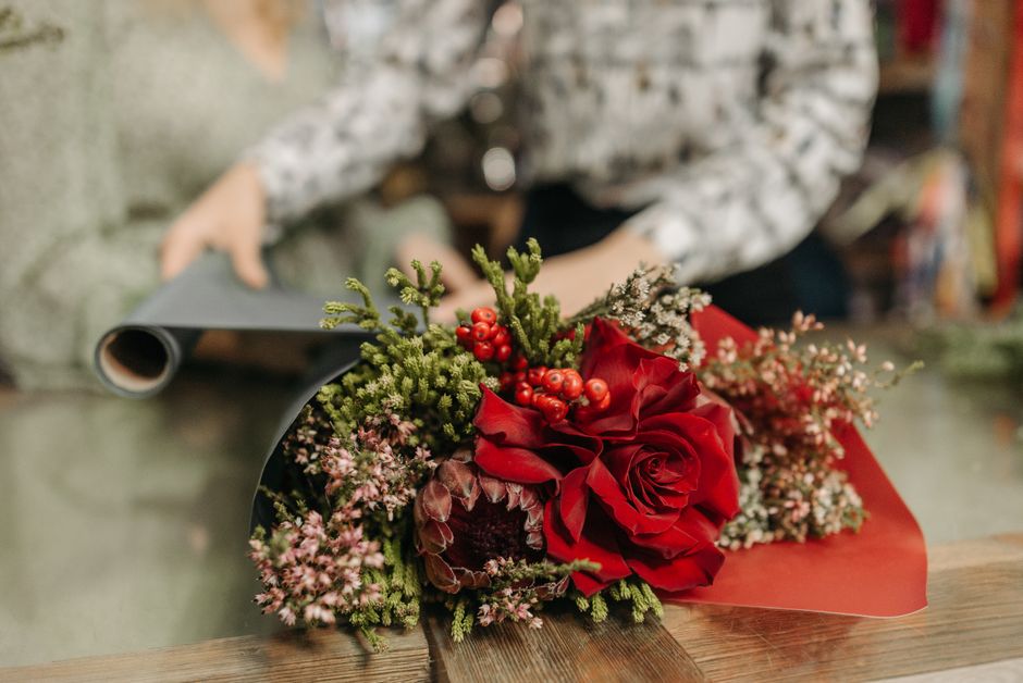 A close-up view of a floral arrangement featuring a large, vibrant red rose surrounded by sprigs of pink and white filler flowers, clusters of red berries, and green foliage, all wrapped in decorative
