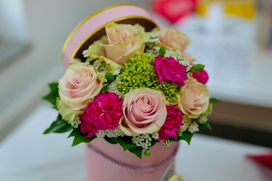 A floral arrangement inside a round, pastel pink cylindrical box with a pink ribbon trim, featuring soft pink roses with slightly open petals, bright fuchsia carnations with ruffled petals, and small
