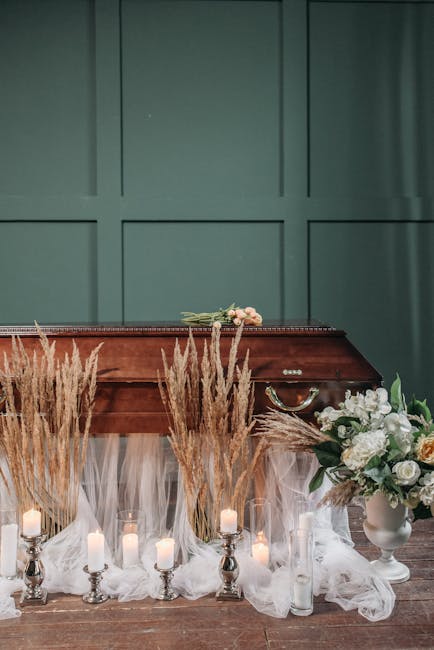 A floral arrangement featuring a bouquet of light pink and white roses placed on a wooden surface in front of a vintage wooden piano. The display includes tall, golden-brown dried grasses and several 