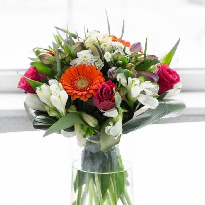 A truly exquisite blend of colour, structure, and heart, this bouquet showcases the best of Rush Green's artisan floristry in a harmonious glass vase display. Gentle peach gerberas, their creamy petals fanning softly, form a delicate ring around the radiant orange centers, reminiscent of sunrise over the local green spaces. Luxurious fuchsia roses, deeply coloured and elegantly shaped, add a romantic counterpoint, while ivory lilies and clusters of pristine spray roses introduce lightness and intricate layering. Broad, glossy aspidistra leaves twist protectively around the bouquet's base, grounding the lively colours much like the historic woodlands anchor Rush Green's sense of place. Wisps of pointed greenery and intricate filler sprays weave through the arrangement, enriching its depth and giving the impression of a blossoming garden freshly gathered for a celebration. Light pours over the bouquet, enhancing every hue from deep ruby to pale blush without ever overwhelming their natural beauty. The imagined fragrance-a gentle mix of fresh rose and subtle lily-lingers, summoning feelings of hope, joy, and togetherness. Designed for gatherings, housewarmings, or thoughtful acts of care along Romford Road and throughout Rush Green, every element of this arrangement expresses optimism and authentic local pride.