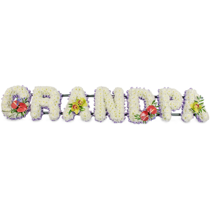 A long, horizontal word tribute reading "GRANDPA" dominates this image, each capital letter a carefully shaped canvas of white blooms set against a clear white background. The letters are completely covered in tightly clustered white chrysanthemums, the blossoms arranged so that their rounded heads almost merge into one continuous surface. This dense coverage creates a soft, textured effect, reminiscent of a quilt or blanket lovingly tucked around treasured memories. Around the base of each letter, a narrow band of light purple or lavender ribbon is visible, adding a delicate outline and giving the design a subtle, cool-toned contrast that remains soothing to the eye. The letters rest securely on dark green bars that run underneath, providing hidden support while keeping the composition elegant and uncluttered. Colourful accent sprays interrupt the expanse of white in a thoughtful rhythm: the 'G' at the beginning and the 'D' near the centre are adorned with rich clusters of deep red anthuriums, their glossy, heart-shaped spathes and bright spadices rising from shiny foliage, suggesting deep affection and strength. The first 'A' and the last 'A' are softened with stems of yellow-green orchids, probably cymbidium, their arching forms and intricate throats bringing an air of grace and gentle optimism. Additional greenery is tucked around these focal flowers, connecting the accents to the chrysanthemum base. The overall feel is one of calm, respectful remembrance, ideal as a personalised funeral tribute for a grandfather in Rush Green, perhaps travelling from a florist near Romford Road to a local chapel. It is a design that speaks quietly yet clearly of love, gratitude, and a life well lived.